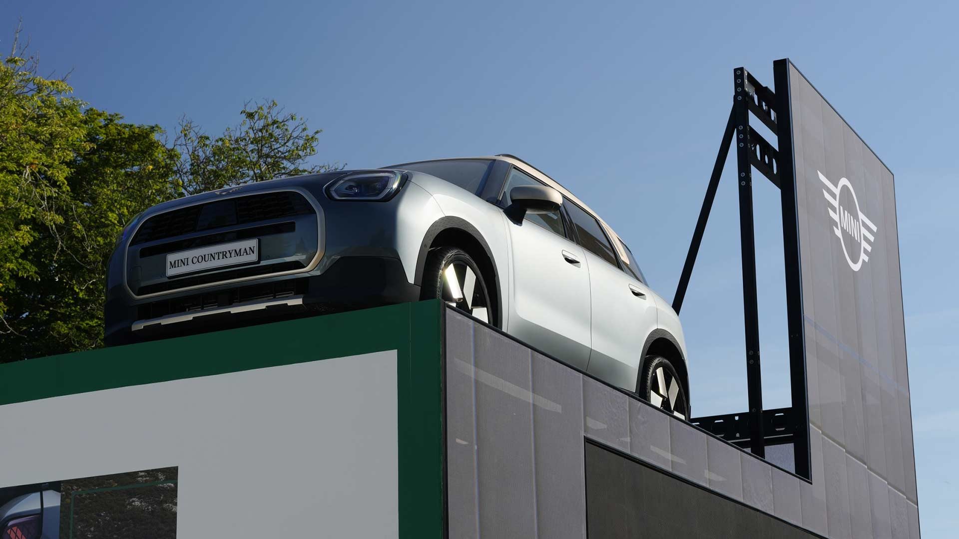 Silver Mini Countryman displayed on a platform against a clear blue sky, surrounded by trees. A large Mini logo is visible on a nearby sign. 